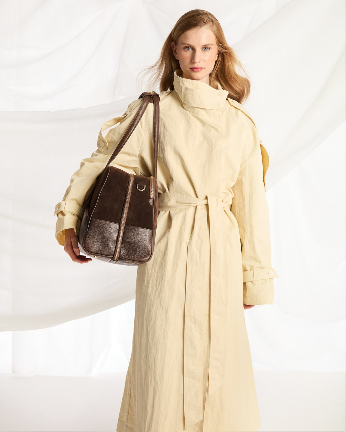 A woman wearing a long cream trench coat and carrying a dark brown leather shoulder bag, posed against a white draped fabric backdrop.