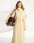 A woman wearing a long cream trench coat and carrying a dark brown leather shoulder bag, posed against a white draped fabric backdrop.