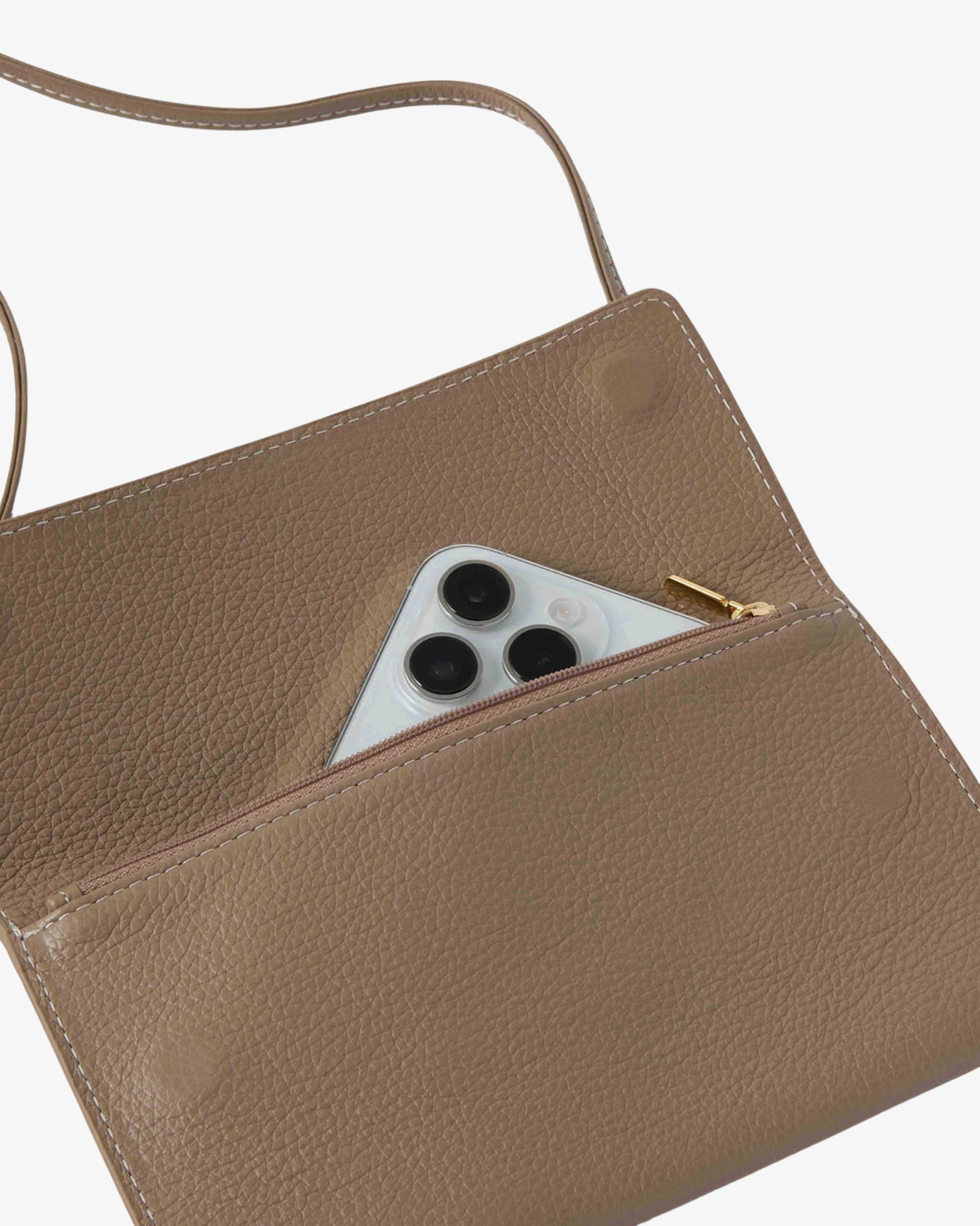 A close-up of a taupe brown pebbled leather crossbody bag with a gold zipper, showing a white smartphone tucked into the front zip pocket against a light gray background.