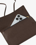 A close-up of a dark brown pebbled leather clutch or pouch with a zippered pocket, shown open with a silver smartphone partially tucked inside. A thin brown strap is visible at the top left against a clean white background.