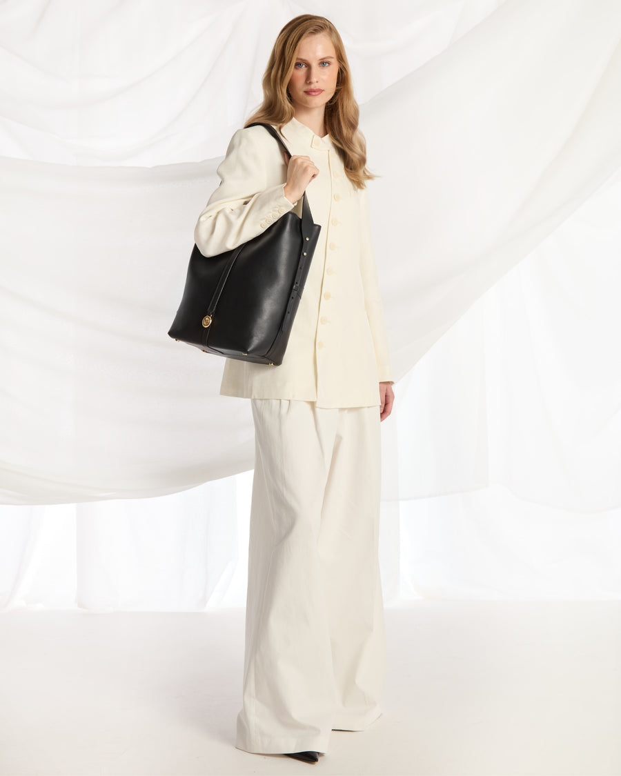 A woman modeling a large black leather shoulder bag with a gold-tone circular logo clasp, styled with a cream-colored blazer and wide-leg trousers against a draped white fabric backdrop.