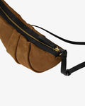 A close-up of a brown suede shoulder bag with pleated detailing, a black leather zipper track, gold-tone hardware, and a black adjustable strap against a light gray background.