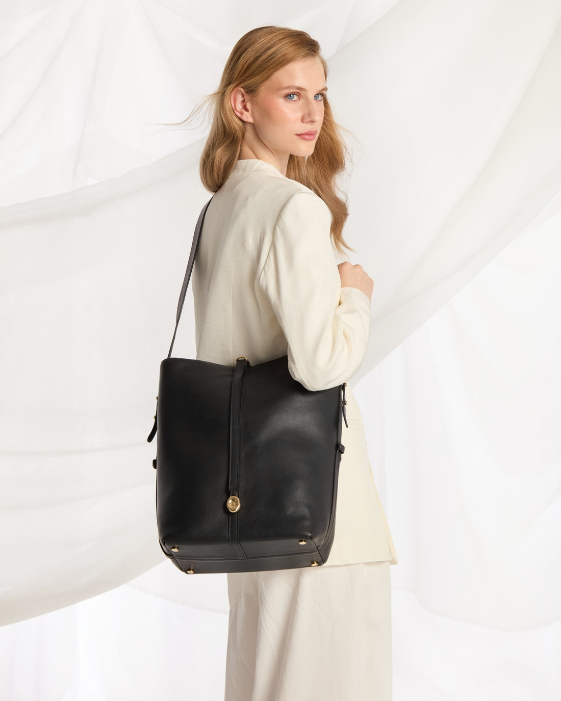 A woman wearing a cream-colored outfit carries a large black leather shoulder bag with gold-tone hardware against a white draped fabric background.