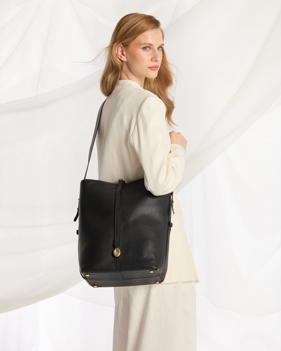 A woman wearing a cream-colored outfit carries a large black leather shoulder bag with gold-tone hardware against a white draped fabric background.
