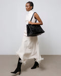 A model in a white textured dress and black boots carries a large black suede shoulder bag against a minimalist studio backdrop.