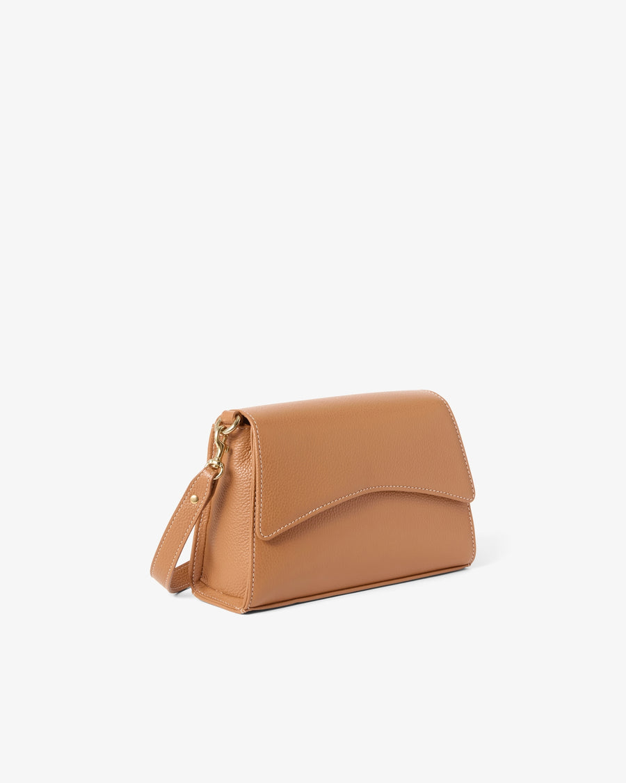 A tan leather crossbody handbag with a curved flap and a matching shoulder strap, shown from a three-quarters angle on a light gray background.