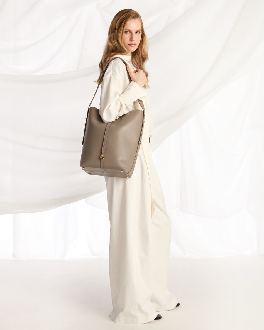 A woman in a cream-colored outfitcarries a large taupe leather shoulder bag against a white draped fabric backdrop.