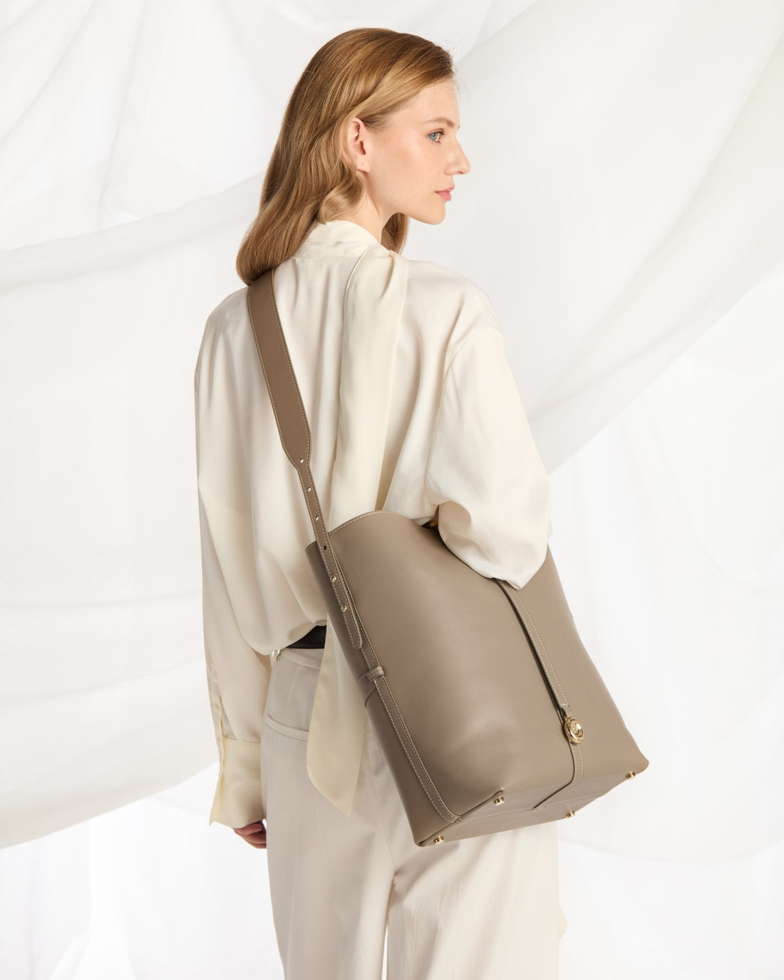 A woman in a cream-colored blouse and trousers is shown from the back and side, carrying a large taupe leather shoulder bag. The background consists of draped white fabric.