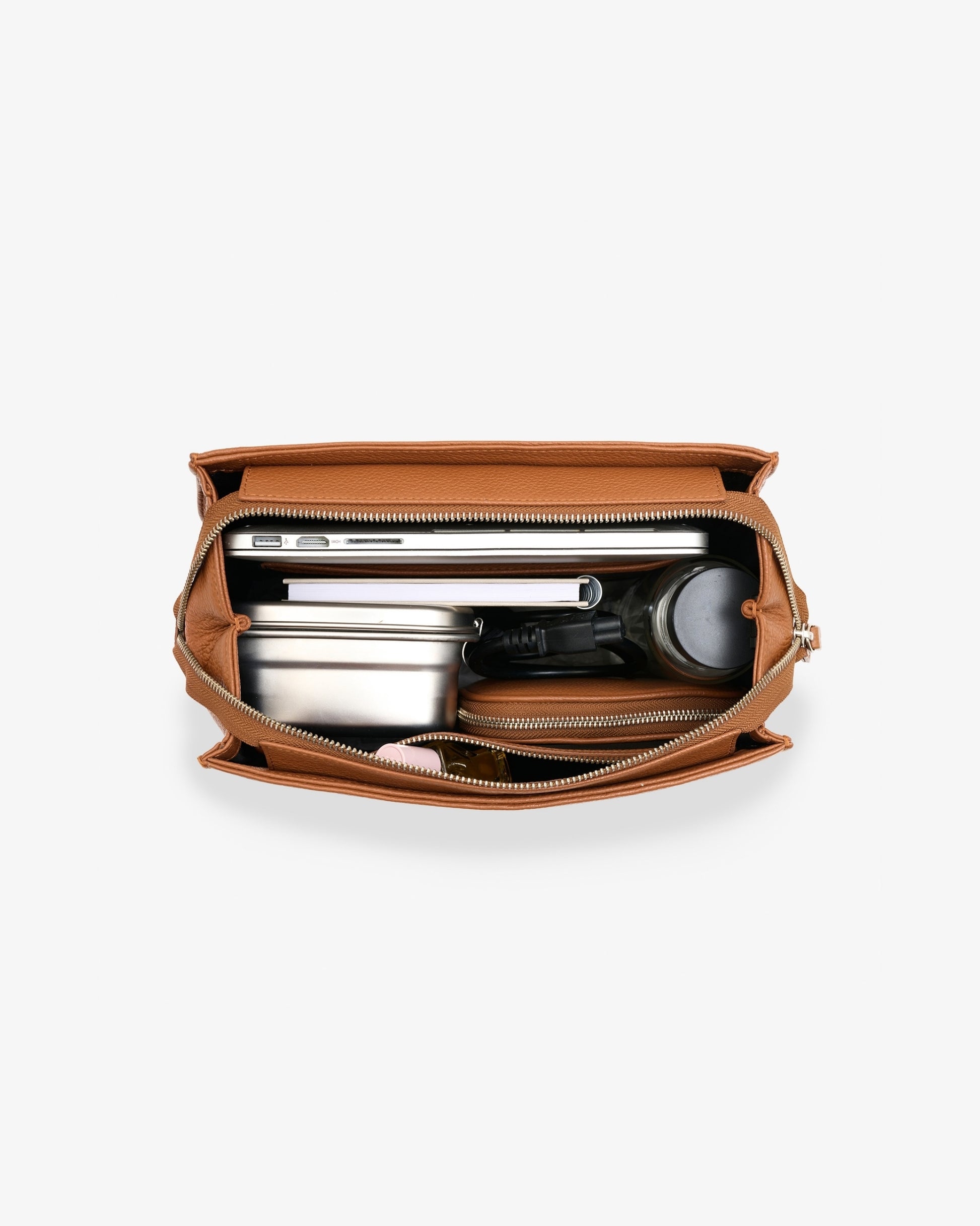 An open tan leather backpack shown from above, revealing its interior compartments filled with a silver laptop, notebooks, a metal tin, a black pen, and a round black item, on a light gray background.