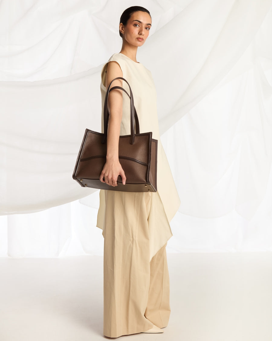 A model in a cream-colored sleeveless dress holds a structured dark brown leather tote bag with double shoulder straps against a white draped fabric backdrop.
