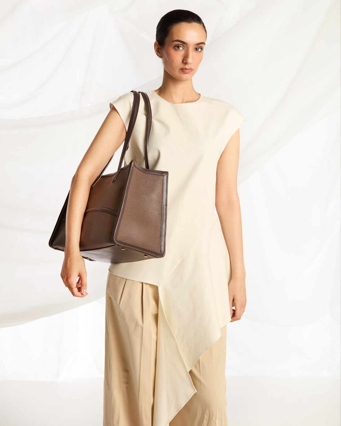 A woman models a large taupe-brown leather tote bag with dark brown shoulder straps, styled with a cream sleeveless top and beige draped trousers against a white draped fabric background.