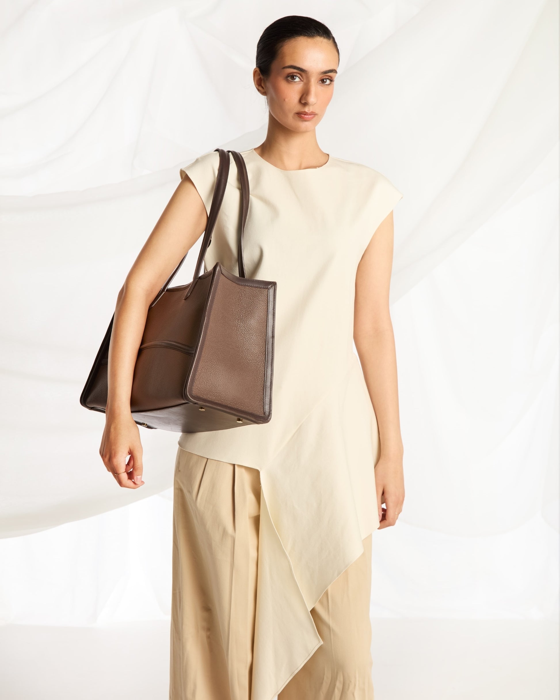A woman models a large taupe-brown leather tote bag with dark brown shoulder straps, styled with a cream sleeveless top and beige draped trousers against a white draped fabric background.