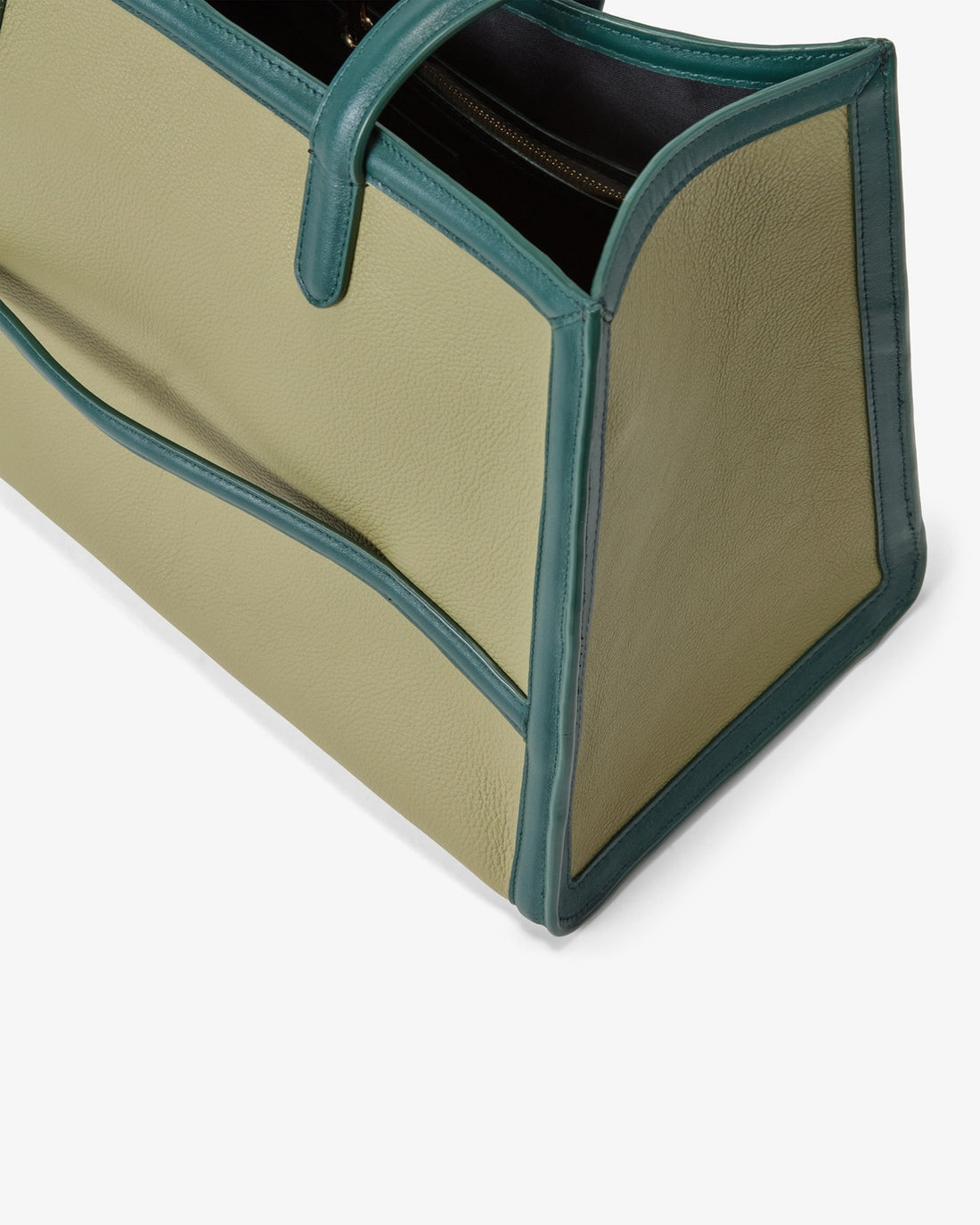 Close-up of a structured tote bag corner showing a light beige pebbled leather body with dark teal-green leather trim and handles as well as a luggage strap, photographed on a clean white background.