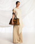 A model in a cream-colored sleeveless dress holds a structured light brown leather tote bag with double shoulder straps against a white draped fabric backdrop.