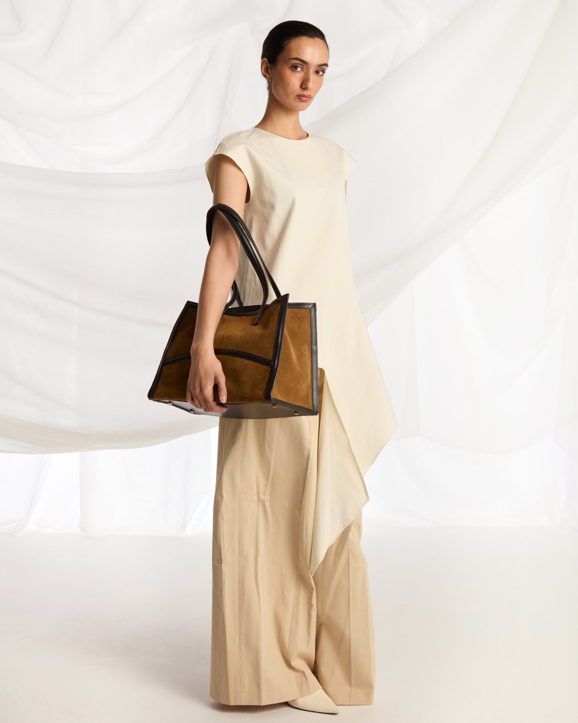 A model in a cream-colored sleeveless dress holds a structured light brown leather tote bag with double shoulder straps against a white draped fabric backdrop.