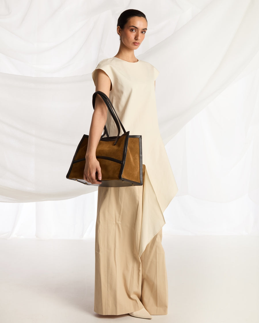 A model in a cream-colored sleeveless dress holds a structured light brown leather tote bag with double shoulder straps against a white draped fabric backdrop.