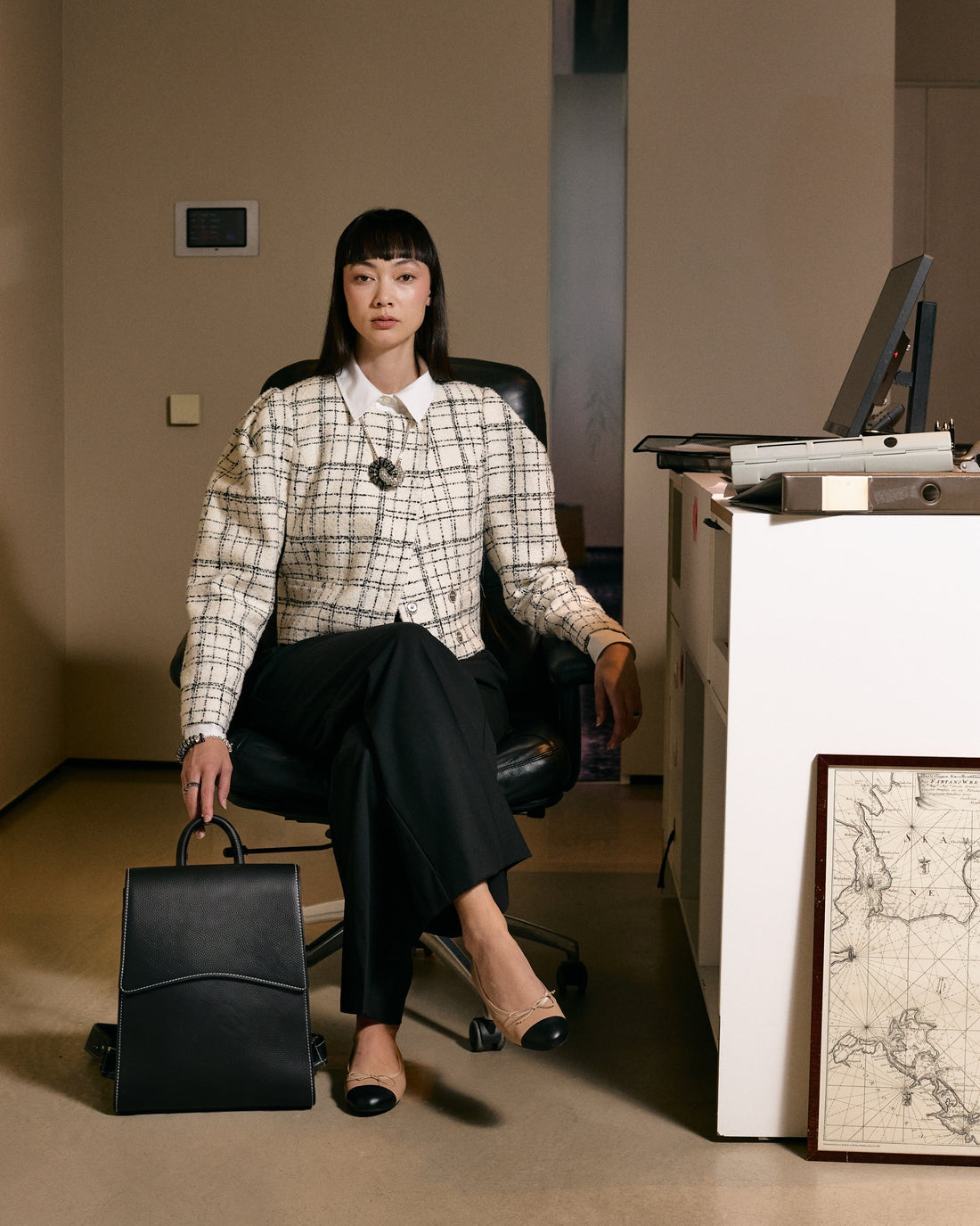 A woman sits in a black office chair in a warm-toned office setting, wearing a cream and black plaid tweed jacket over a white collared shirt, black trousers, and beige heels. She holds a black leather briefcase.