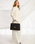 A woman wearing a cream-colored long-sleeve coat and wide-leg trousers holds a structured black handbag with gold-tone hardware against a draped white fabric backdrop.