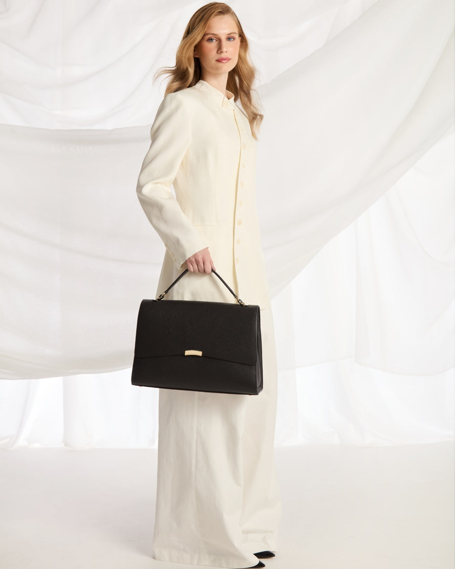 A woman wearing a cream-colored long-sleeve coat and wide-leg trousers holds a structured black handbag with gold-tone hardware against a draped white fabric backdrop.