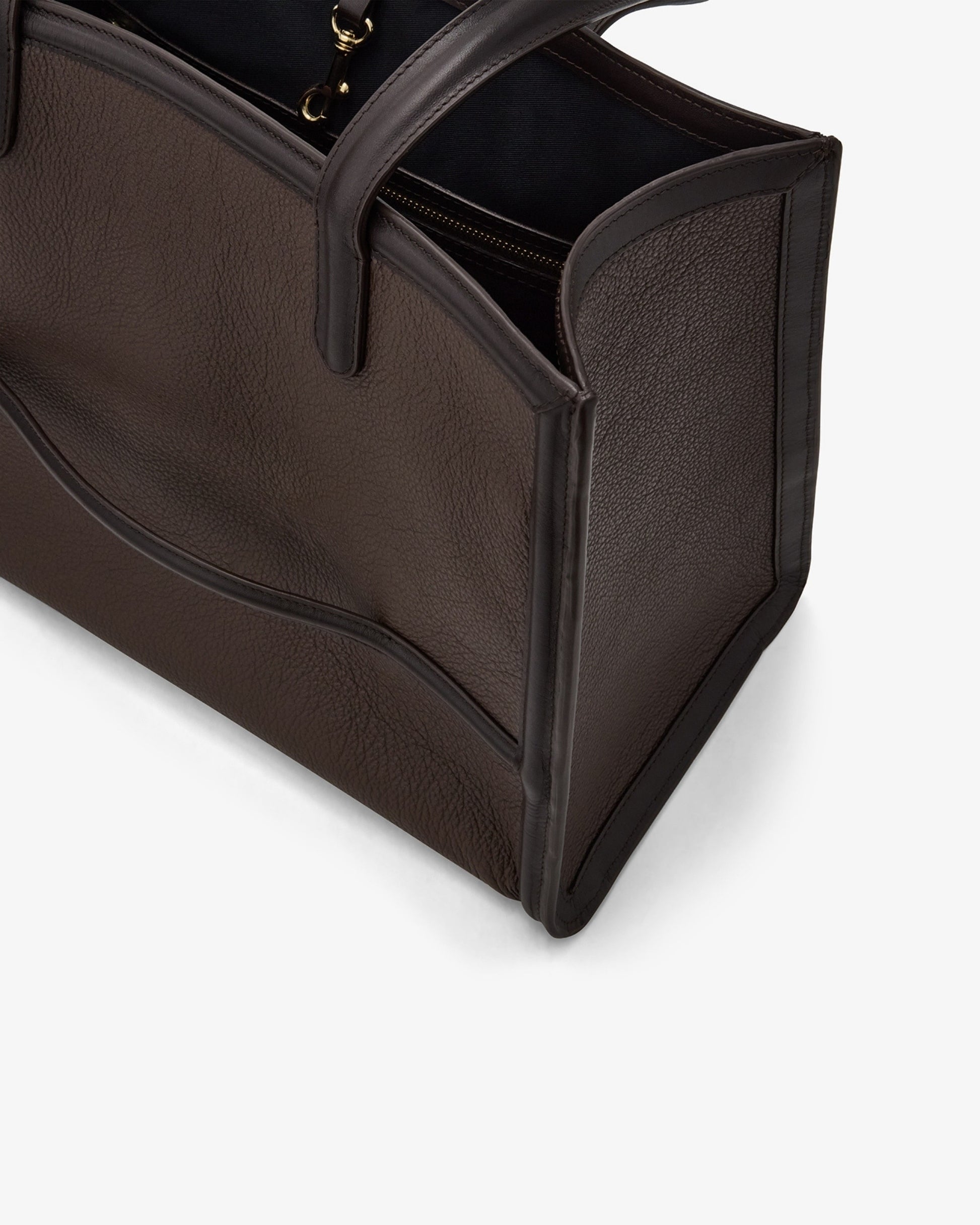 Close-up of a dark brown pebbled leather tote bag with black trim and handles, showing the interior zipper and corner details on a light gray background.