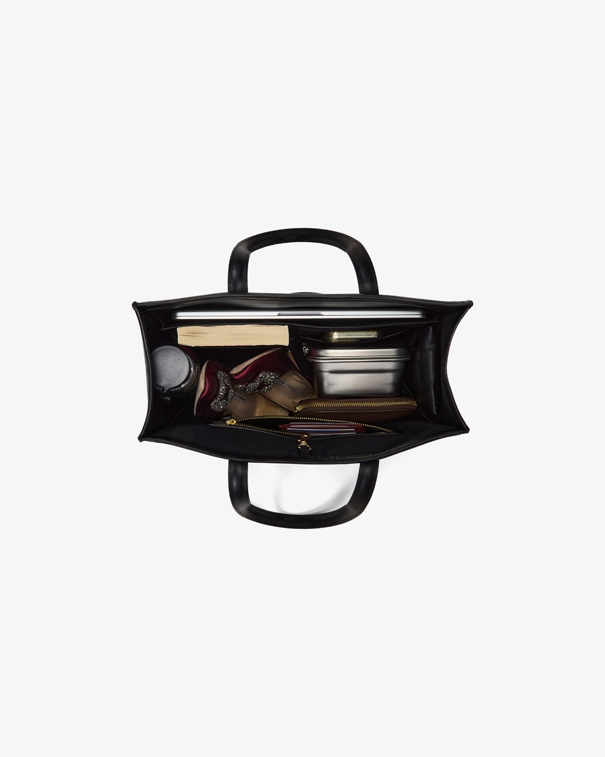 A black leather bag shown from a top-down view, open to reveal a packed interior with a laptop, books, a bottle, and other travel items, set against a plain light gray background.