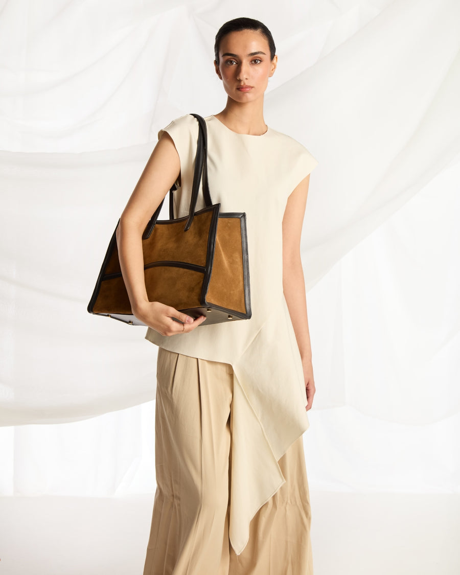 A woman models a large taupe-brown leather tote bag with light brown shoulder straps, styled with a cream sleeveless top and beige draped trousers against a white draped fabric background.