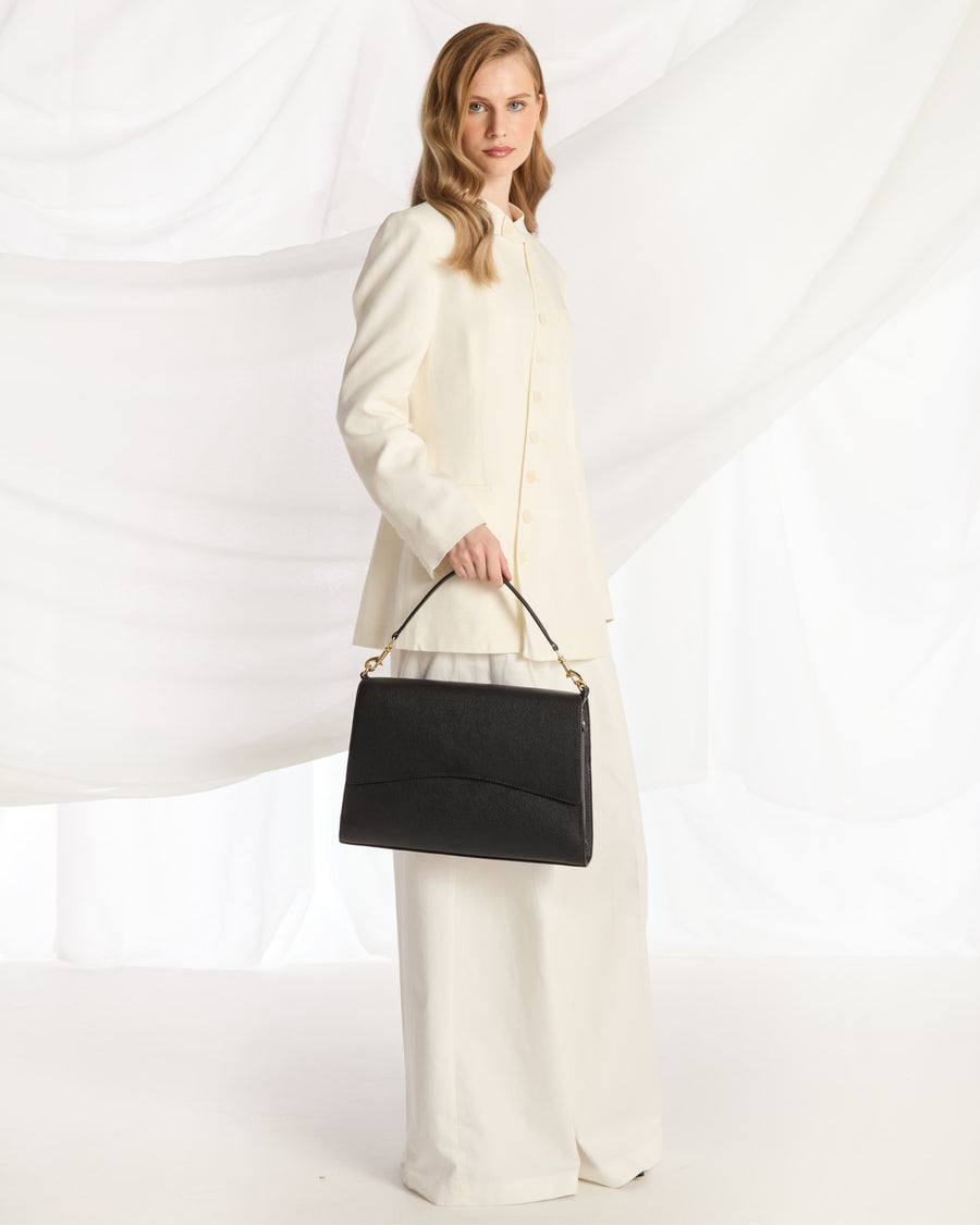 A woman wearing a cream-colored long-sleeve coat and wide-leg trousers holds a structured black handbag with gold-tone hardware against a draped white fabric backdrop.