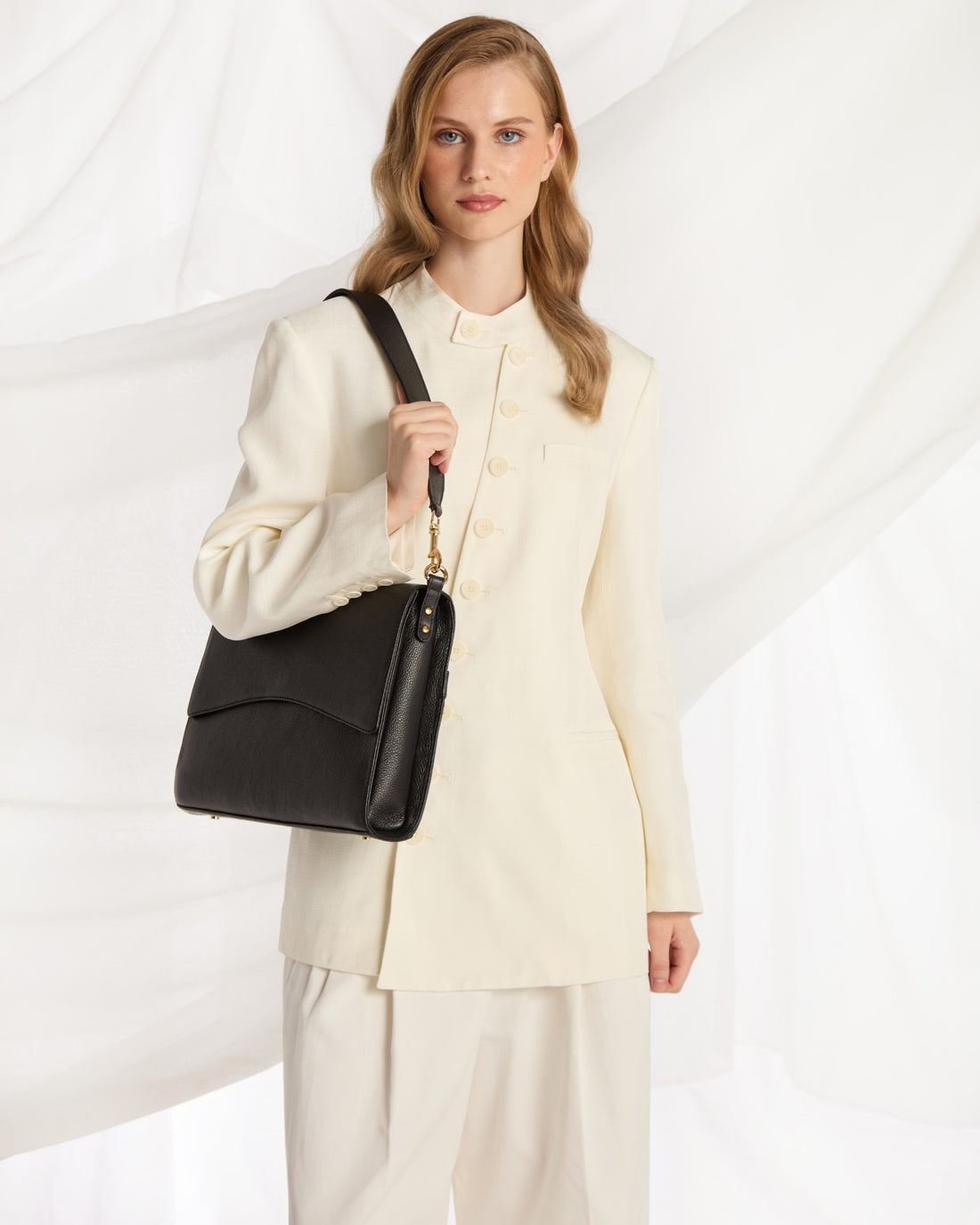 A woman wearing a cream-colored tailored suit and holding a black leather shoulder bag with a gold chain detail, posed against a white draped fabric background.