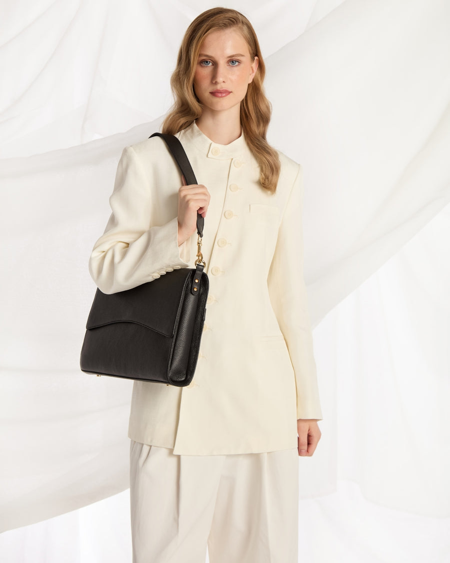 A woman wearing a cream-colored tailored suit and holding a black leather shoulder bag with a gold chain detail, posed against a white draped fabric background.