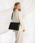 A woman in a cream-colored blazer and trousers carries a black leather shoulder bag with a curved front flap and gold-tone chain details, posed against a white draped fabric background.