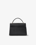 A black leather top-handle handbag with a luggage strap on the bag, shown against a plain white background.