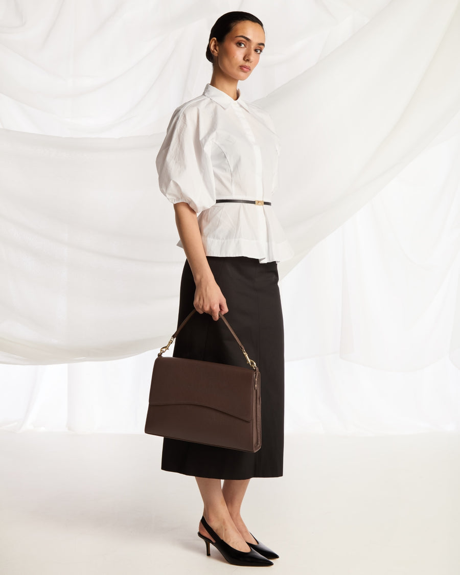 A woman stands in a studio wearing a white puff-sleeve blouse, a black midi skirt, and black heels, while holding a structured dark brown handbag with a gold chain strap.