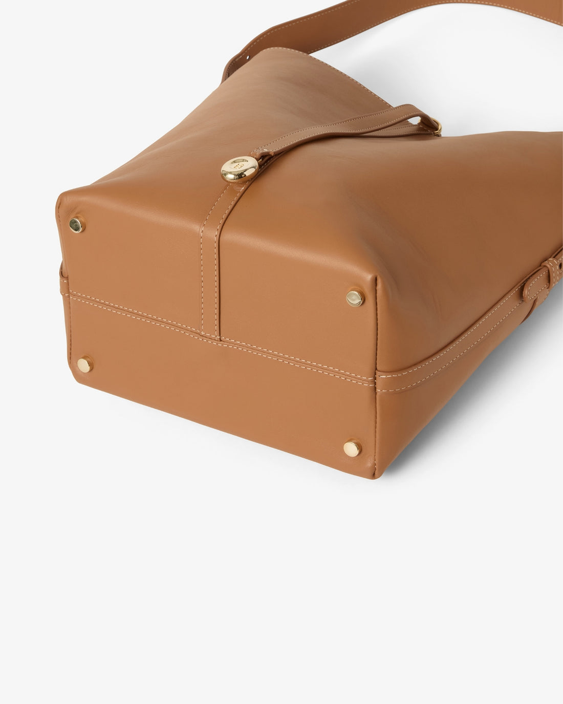 A close-up of a tan leather shoulder bag showing the bottom corner, gold-tone rivets, and a strap closure with a round gold clasp, set against a clean white background.