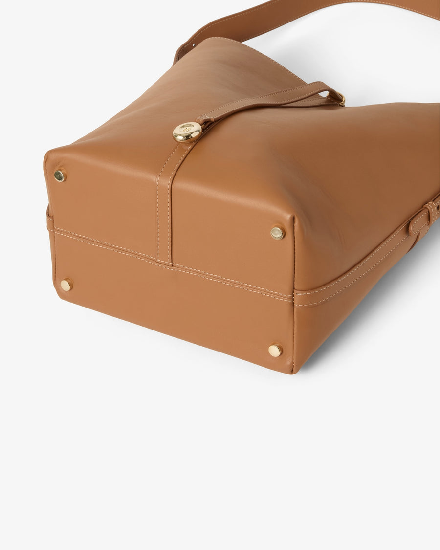 A close-up of a tan leather shoulder bag showing the bottom corner, gold-tone rivets, and a strap closure with a round gold clasp, set against a clean white background.