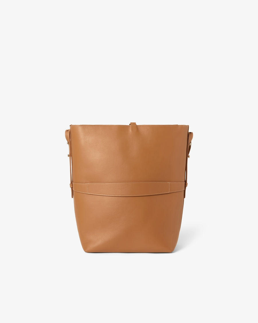 A tan leather bucket bag with a structured rectangular silhouette, a horizontal luggage strap, and a matching leather shoulder strap, shown against a plain white background.