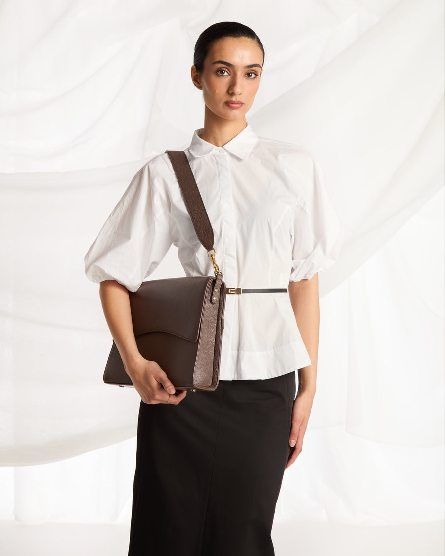 A woman wearing a white puff-sleeve blouse and black skirt carries a brown leather shoulder bag with a matching strap and gold-tone hardware against a draped white fabric backdrop.