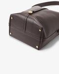A close-up of a dark brown leather shoulder bag showing the bottom corner, gold-tone hardware, and strap attachment on a light gray background.