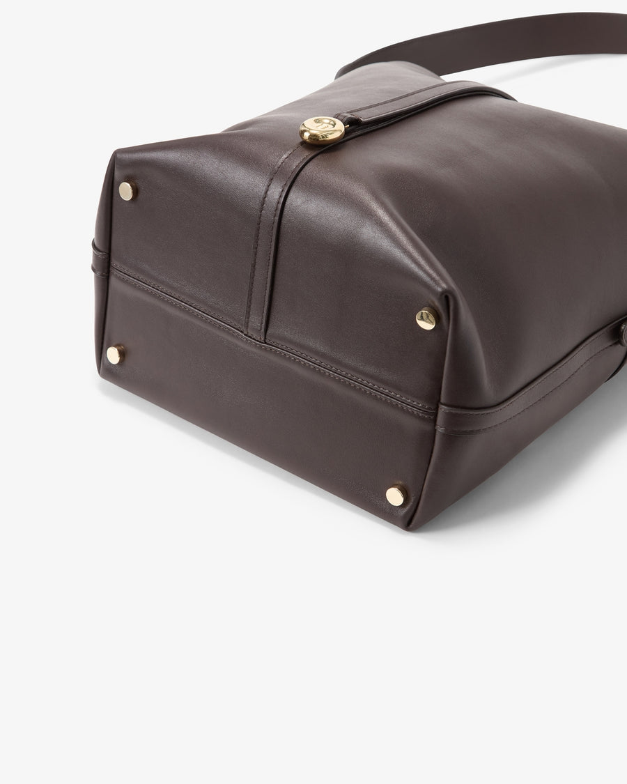 A close-up of a dark brown leather shoulder bag showing the bottom corner, gold-tone hardware, and strap attachment on a light gray background.