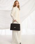 A woman wearing a long cream-colored coat and wide-leg trousers holds a structured black leather handbag with a top handle against a draped white fabric backdrop.