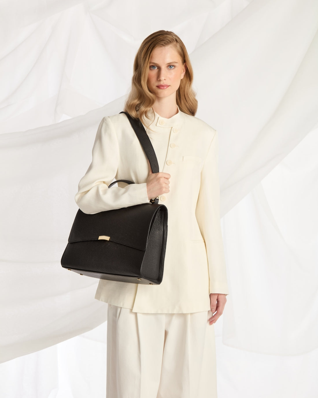 A woman wearing a cream-colored tailored suit and holding a black leather shoulder bag with a strap, posed against a white draped fabric background.
