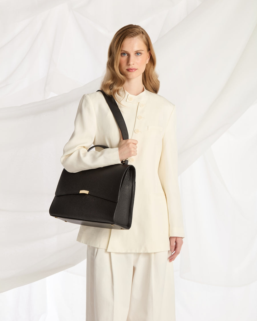 A woman wearing a cream-colored tailored suit and holding a black leather shoulder bag with a strap, posed against a white draped fabric background.
