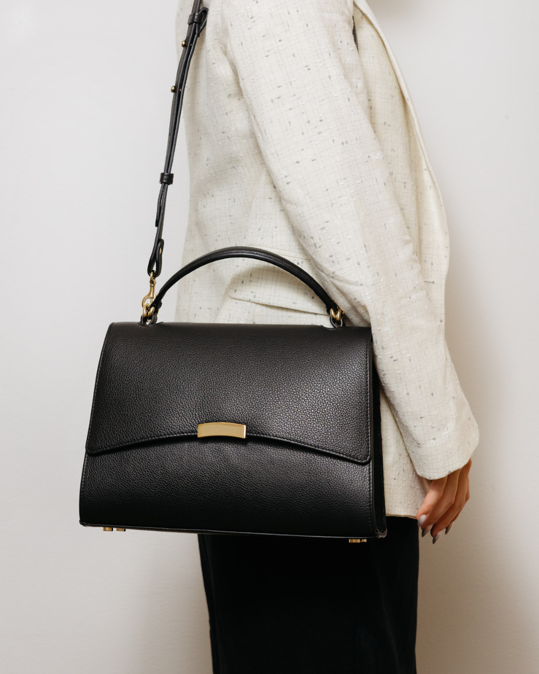 Black handbag held by a person wearing a white sweater and black pants on a light background