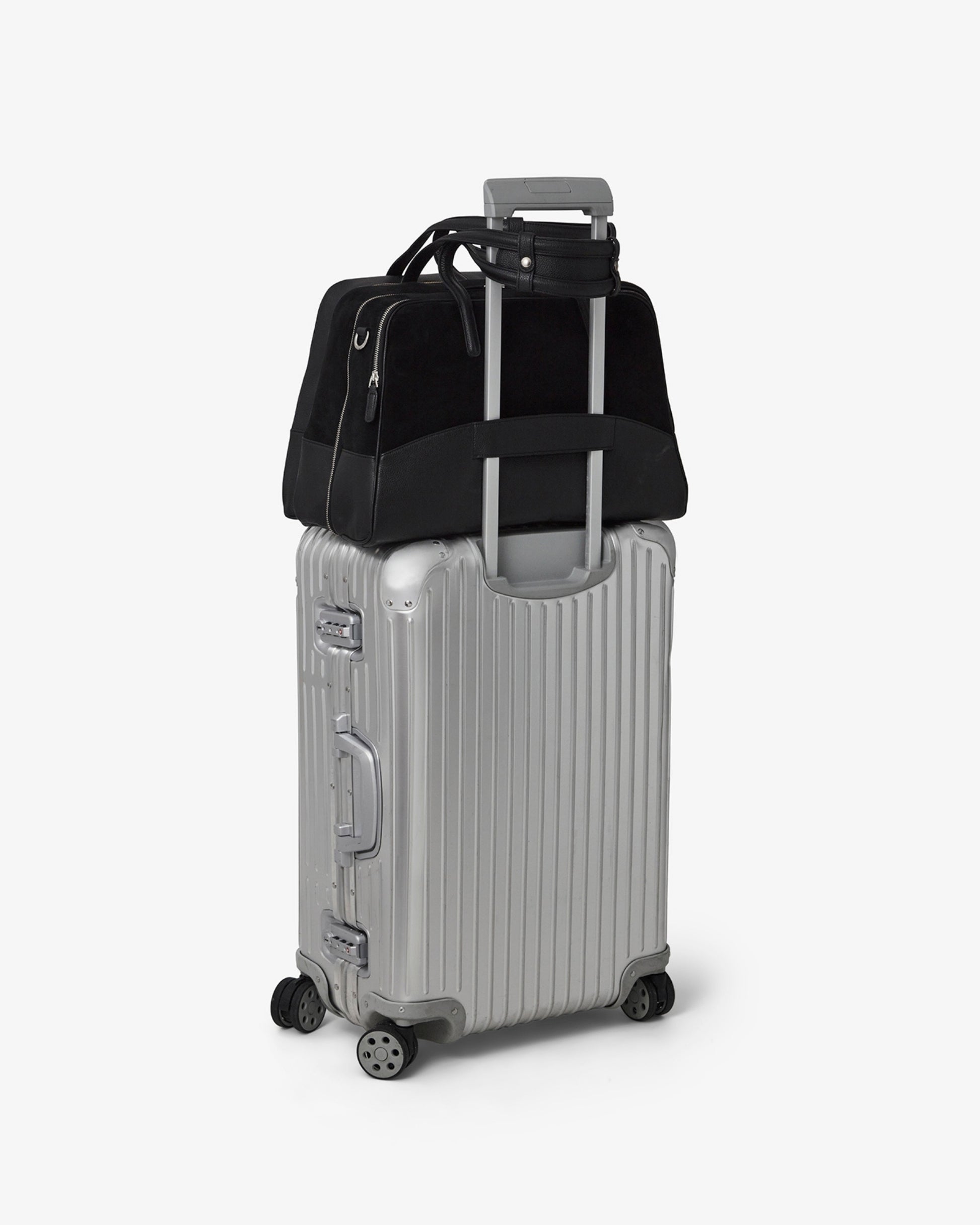 A silver hard-shell spinner suitcase with a telescoping handle extended, carrying a black duffel bag on top, photographed on a clean light gray studio background.
