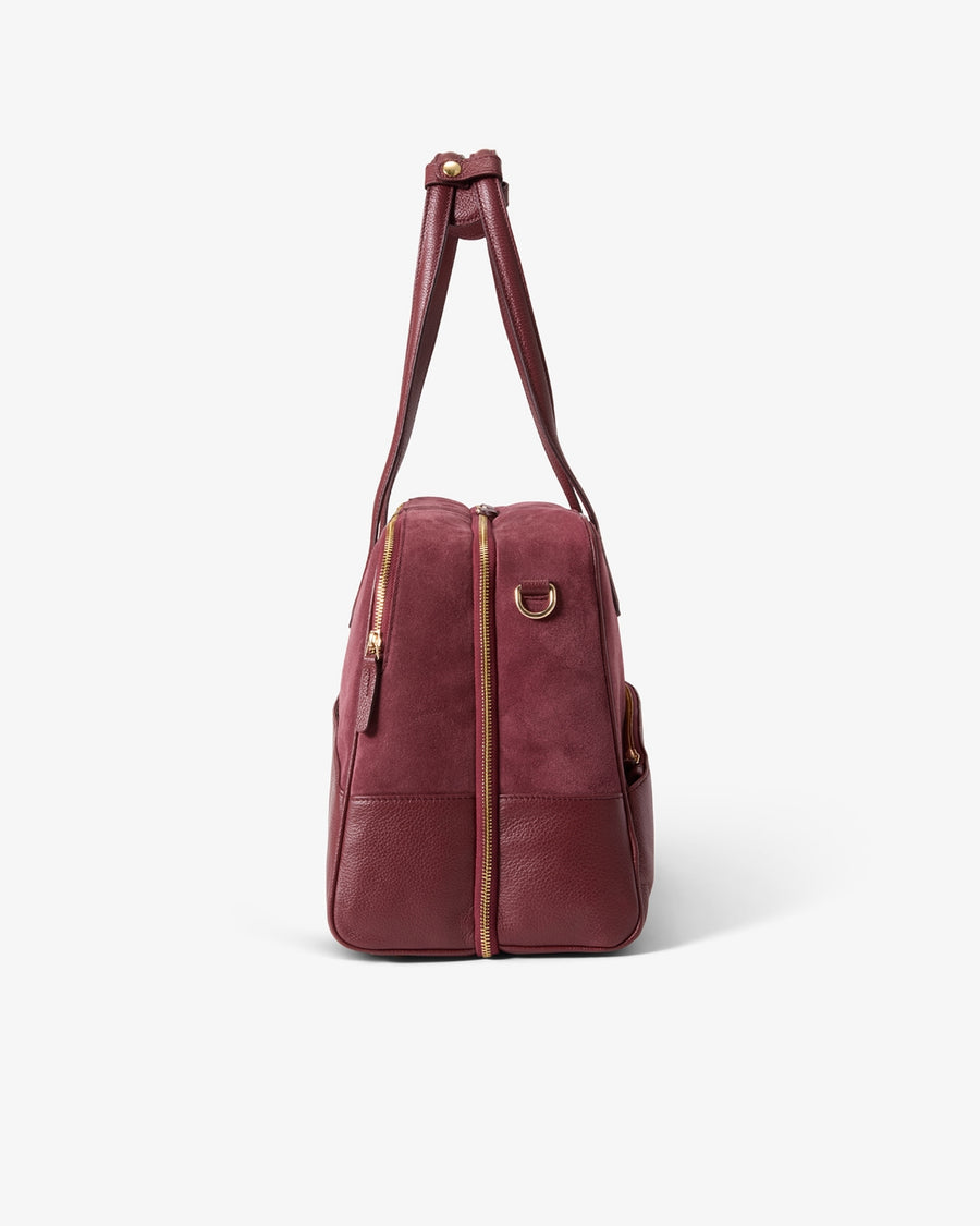A burgundy leather duffel bag with gold-tone hardware, shown from the side on a clean white background.