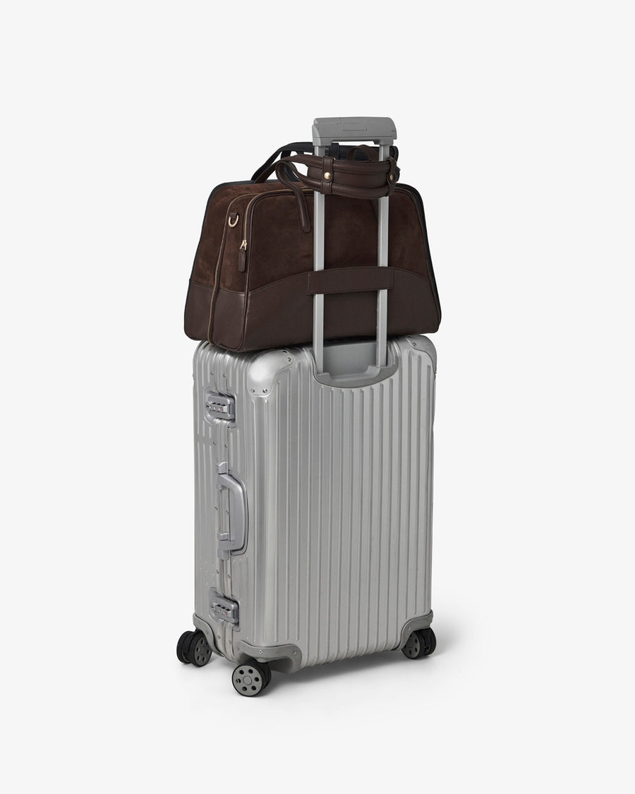 A dark brown leather carry-on bag with a telescoping handle, resting on top of a silver ribbed hard-shell suitcase against a light gray background.