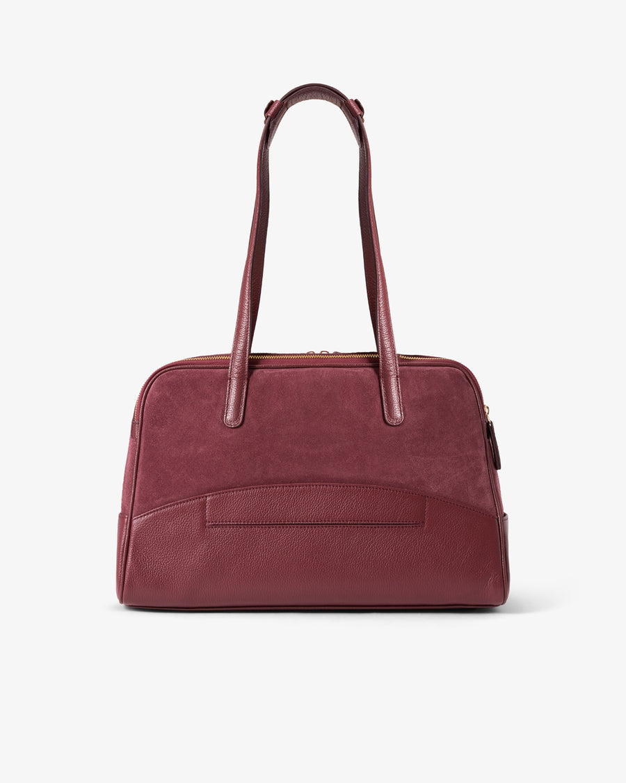 A burgundy leather handbag with dual top handles and a front slip pocket, as well as a luggage strap, photographed against a clean white background.