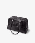 A black crocodile-embossed leather duffel bag with dual top handles and a zippered top closure, photographed on a clean white background.