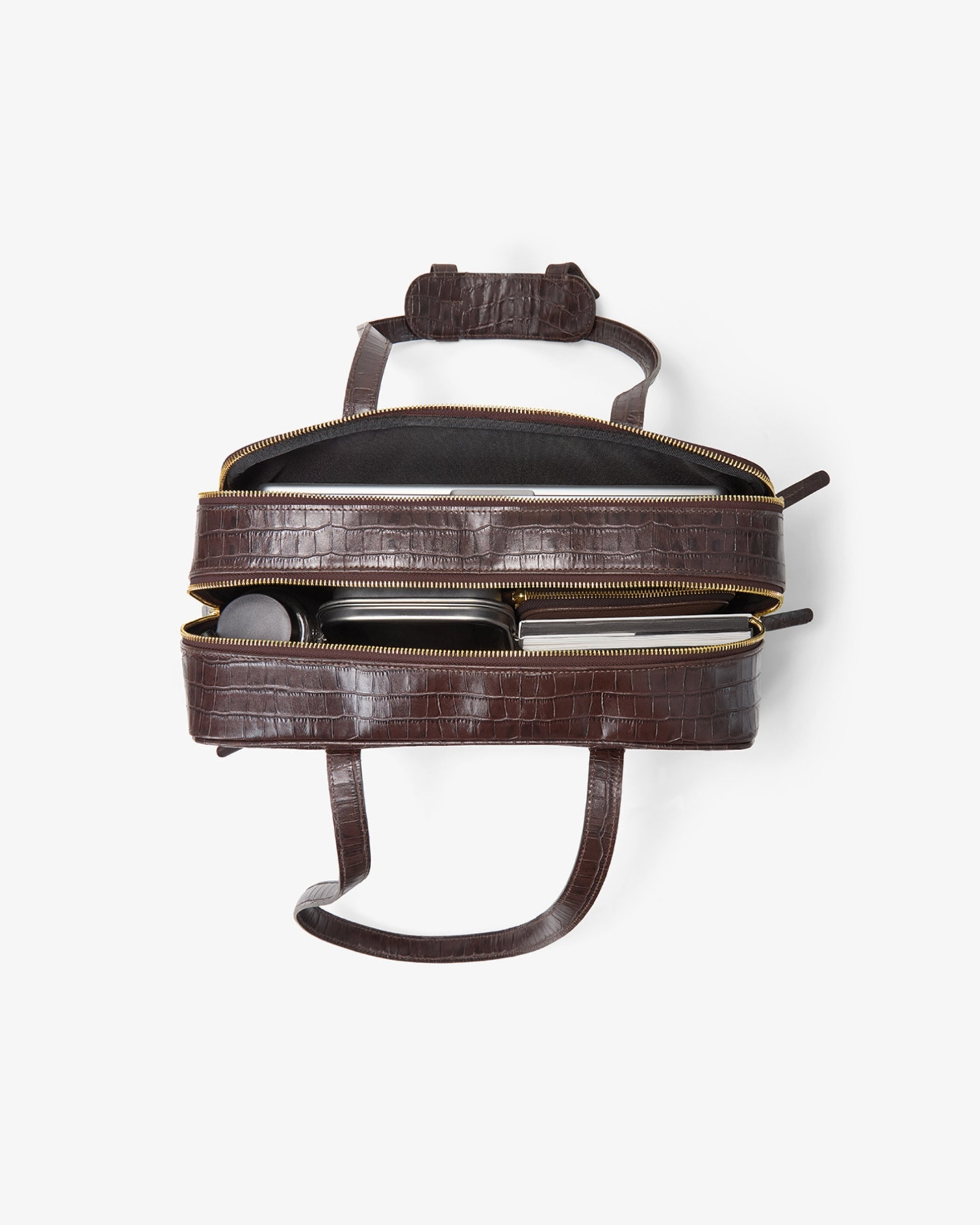 A dark brown crocodile-embossed zippered leather bag shown open from a top-down view, revealing its interior and contents, on a clean white background.