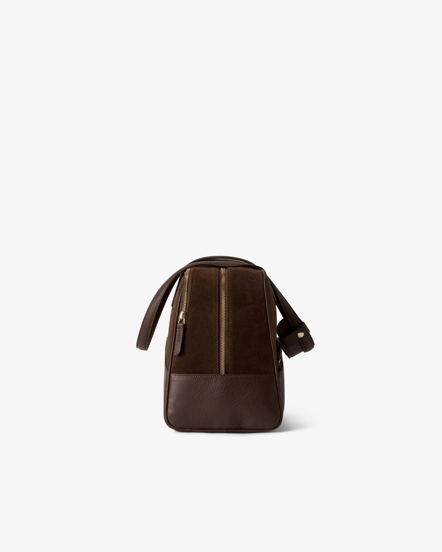 A dark brown leather duffel bag with a vertical zippered compartment and two top handle, shown from the back on a light gray background.