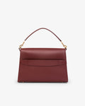 A structured burgundy leather handbag with a top handle, gold-tone hardware, and a luggage strap, shown against a plain light gray background.
